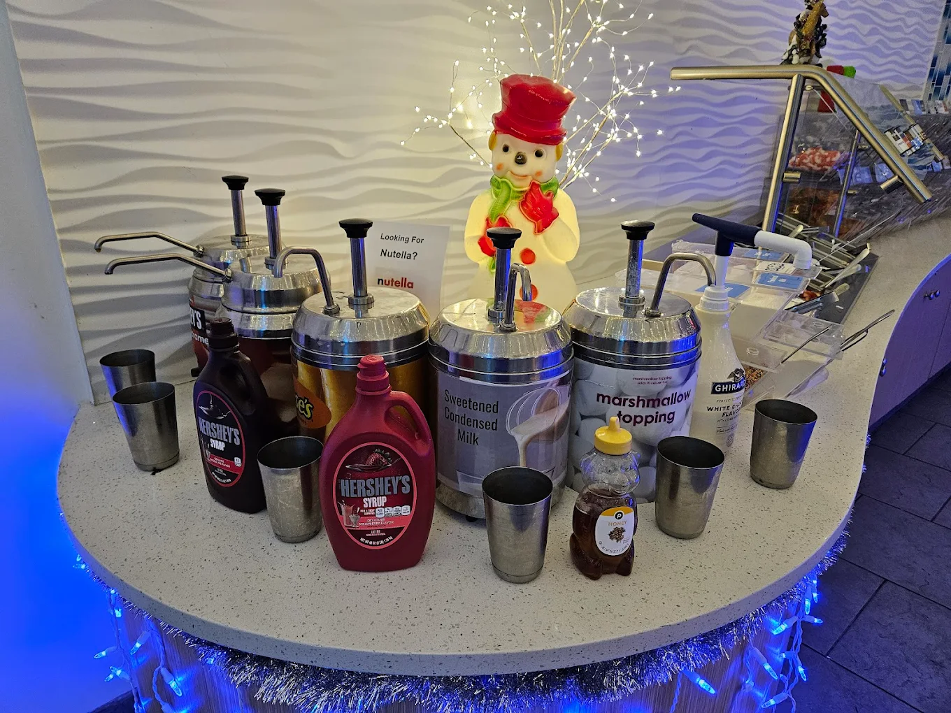 Frosty Froyo toppings station featuring circular counter with blue LED lighting, various syrups including Hershey's and Nutella, marshmallow topping, and festive snowman decoration in Champions Gate
